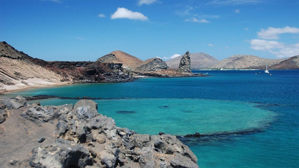 Rocky coastline with clear turquoise water, sandy beaches, and distant hills under a blue sky with a few clouds. Sailboats are visible on the right side of the image.
