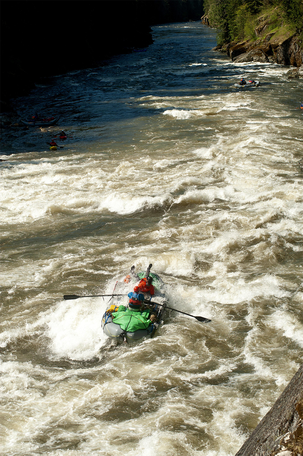 A group of people in a green inflatable raft navigate whitewater rapids on a river, surrounded by rocky banks and trees.