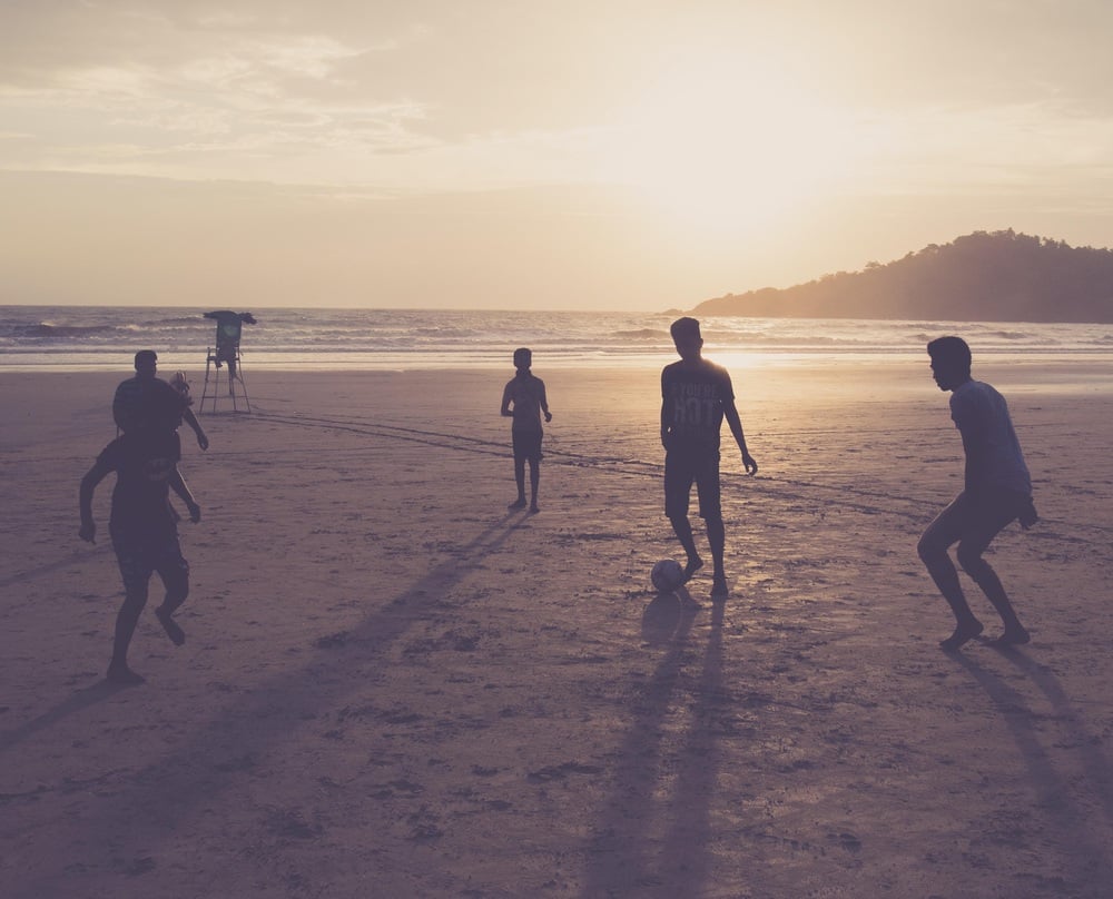 Five people play soccer on a sandy beach at sunset, casting long shadows. The ocean and a hilly landscape are visible in the background.