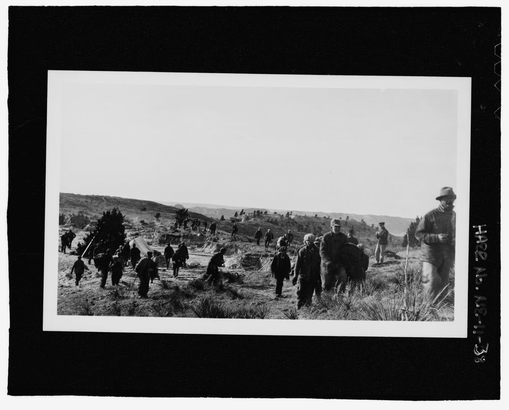 A group of people walk across a hilly, grassy landscape carrying bundled belongings; some figures appear to be leading animals. The scene appears historic and somber.