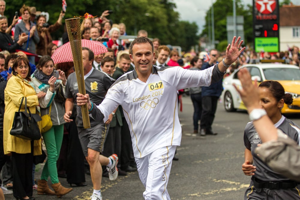 A man in a white London 2012 Olympic uniform carries the Olympic torch, smiling and waving to a cheering crowd on a street. People watch and take photos as he runs.