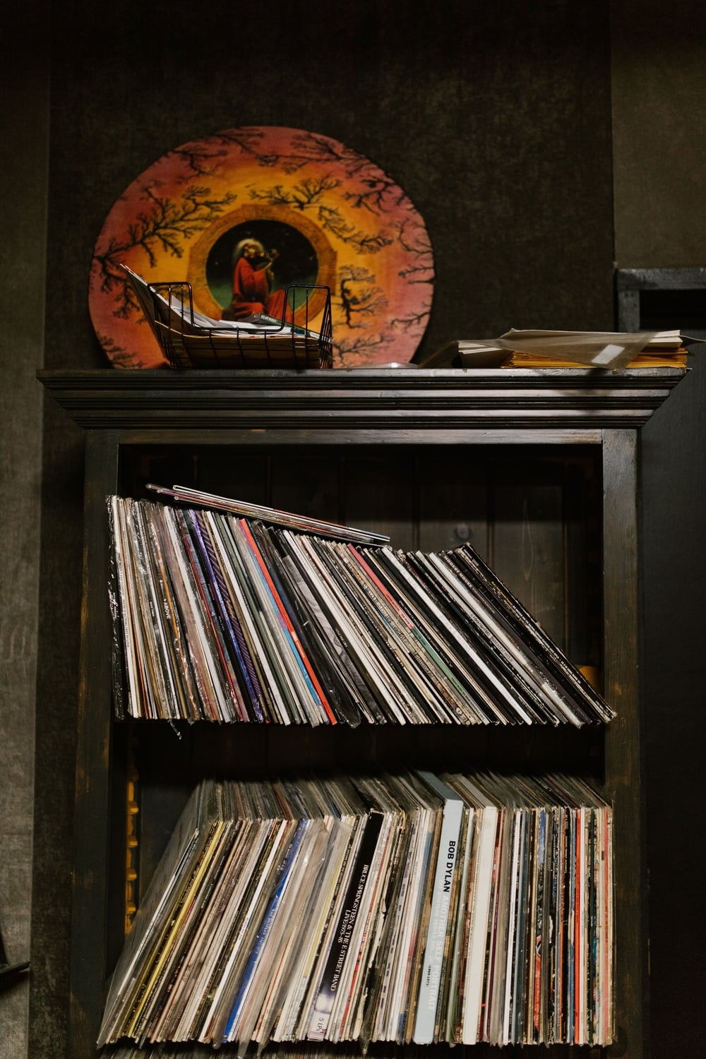 A shelf holds two rows of vinyl records, with a large decorative plate featuring a painted figure and trees resting in a wire stand on top.