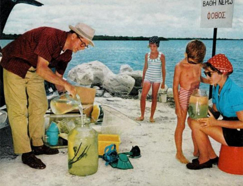 A man pours liquid into a large jar while a woman and two children in swimsuits watch by a lakeshore. There are rocks, water, and a Greek sign in the background.
