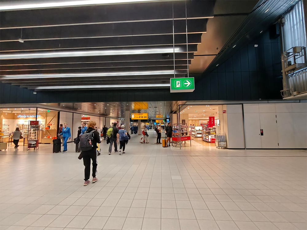 People walk through a spacious, well-lit airport terminal with shops on both sides and green exit signs hanging from the ceiling.