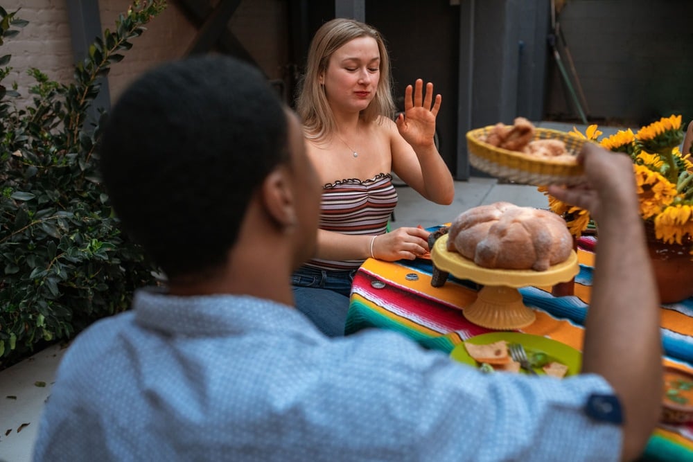 A woman gestures “no” with her hand while a man offers her bread at a colorful outdoor table decorated with sunflowers and pastries.