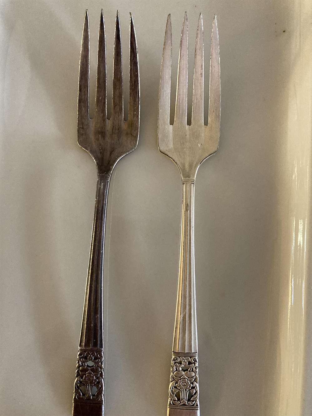 Two metal forks with ornate handles are placed side by side on a white surface. The fork on the left is tarnished, while the fork on the right is clean and shiny.