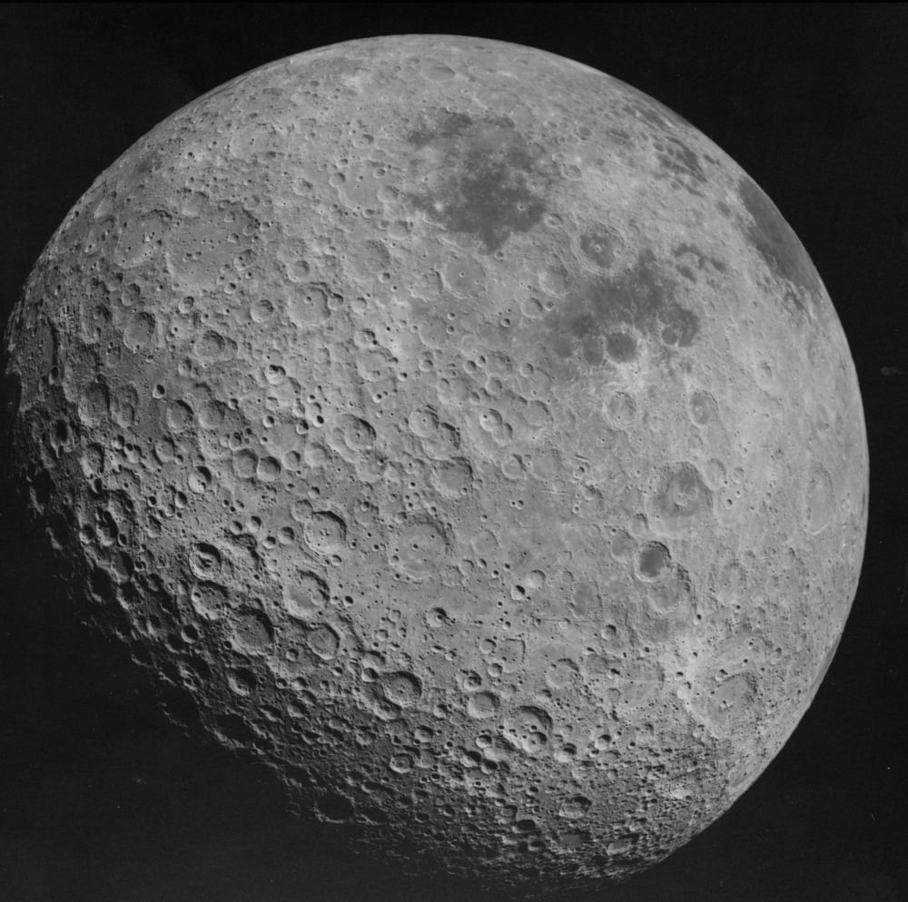 A detailed black and white photograph of the moon’s surface, showing numerous craters and rough terrain, with some areas appearing smoother. The image captures the moon against a completely dark background.