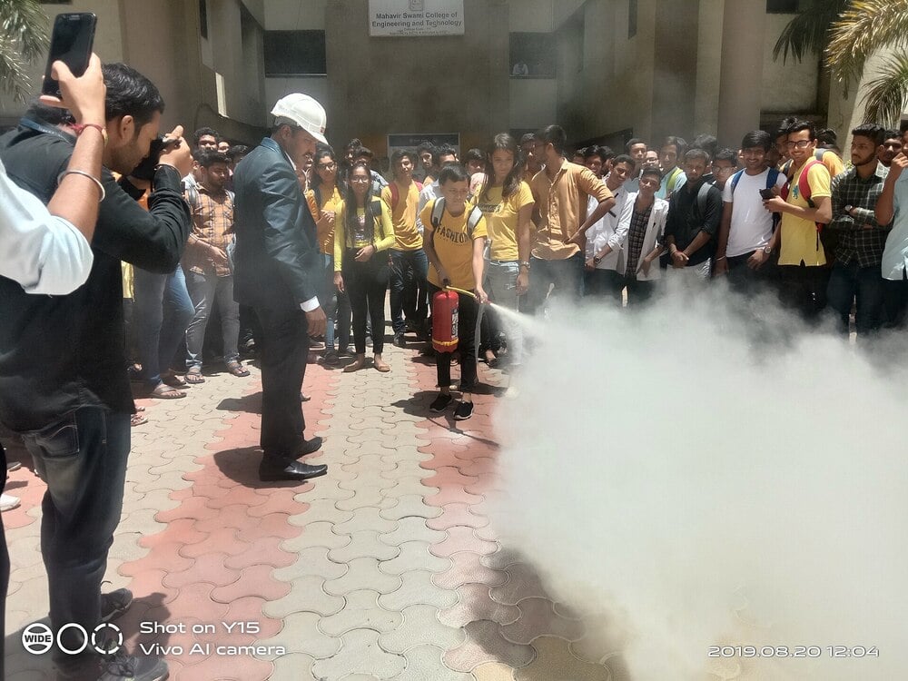 A crowd watches as a person uses a fire extinguisher, producing thick white smoke, during a safety demonstration outside a building in daylight.