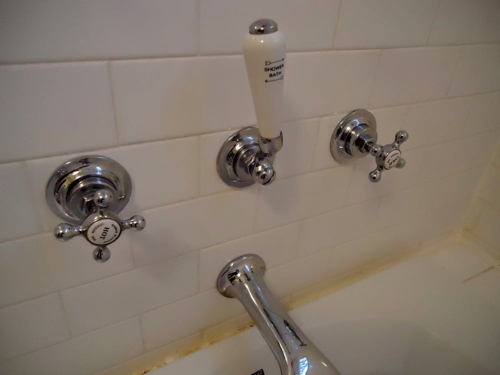 Three chrome faucet handles and a spout are mounted on white tile. The center handle is vertical with a white grip, while the left and right handles are cross-shaped.