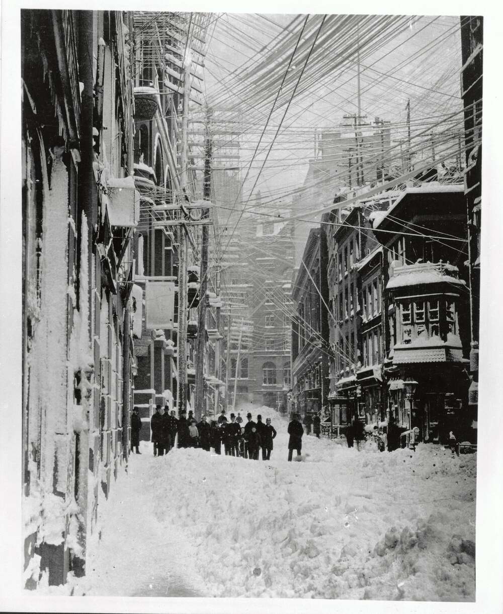A group of people stand in a snow-covered city street lined with tall buildings and dense overhead power lines during a winter storm in the late 1800s.