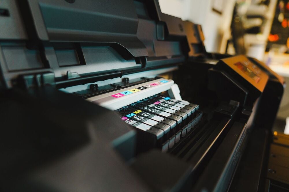 Close-up of an open printer showing multiple ink cartridges in different colors, ready for printing. The focus is on the cartridges, with the surrounding printer parts slightly blurred.