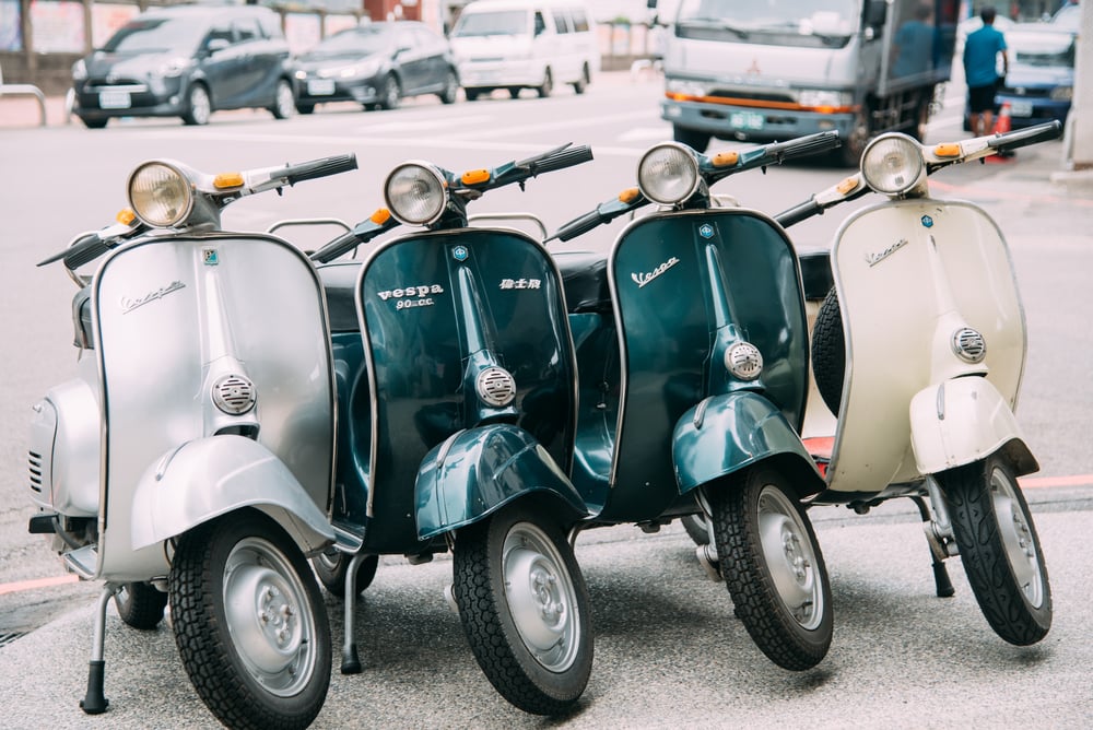 Four vintage Vespa scooters, in silver, blue, and cream, are parked side by side on a city street with vehicles visible in the background.
