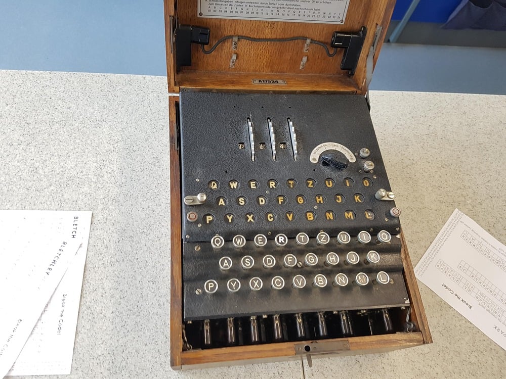 A vintage Enigma machine with keys, rotors, and a plugboard, used for code-making during World War II, sits on a light-colored tabletop with papers nearby.