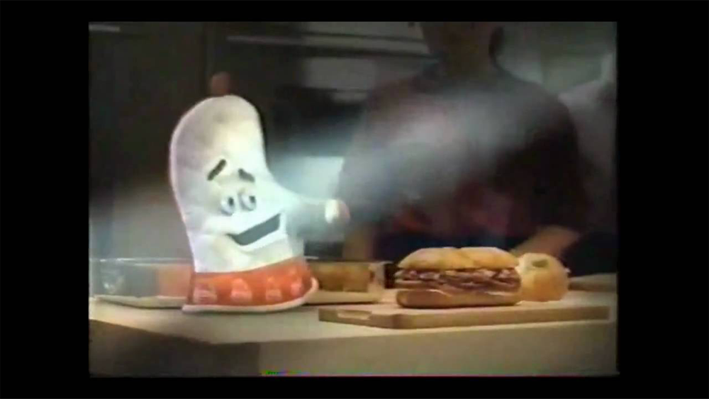 A smiling animated oven mitt gestures toward a sandwich on a kitchen counter, with two people blurred in the background.