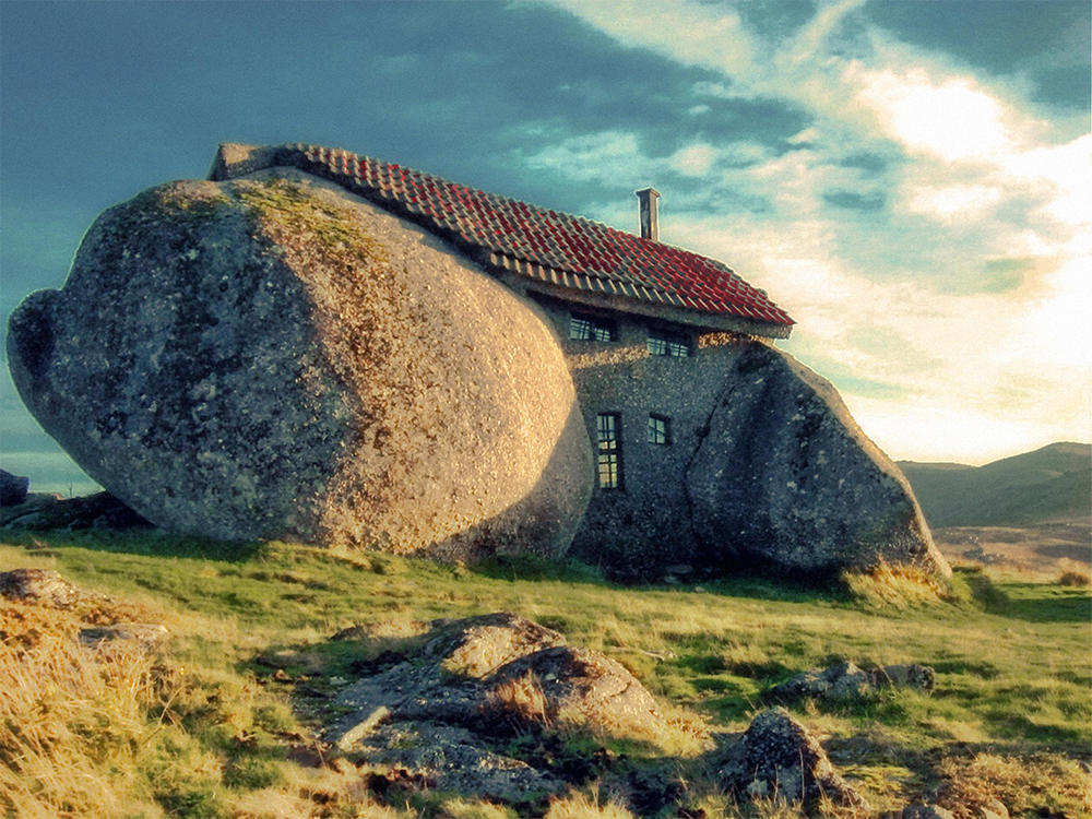 A small stone house with a red-tiled roof is built between and beneath large boulders on a grassy hillside under a partly cloudy sky.