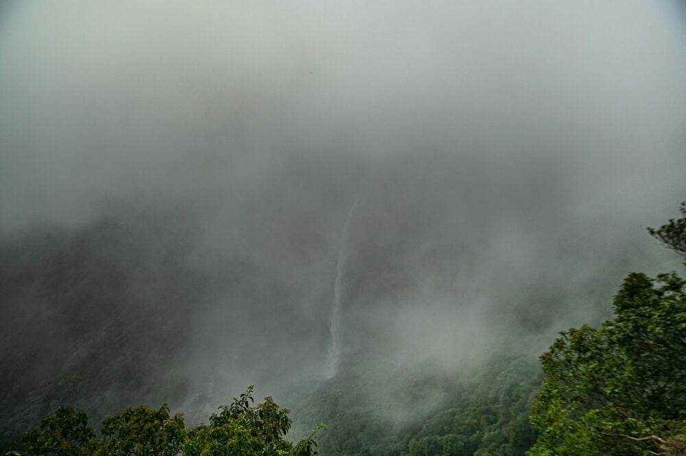 A tall, narrow waterfall flows down a green, forested mountainside, partially obscured by dense fog and mist.