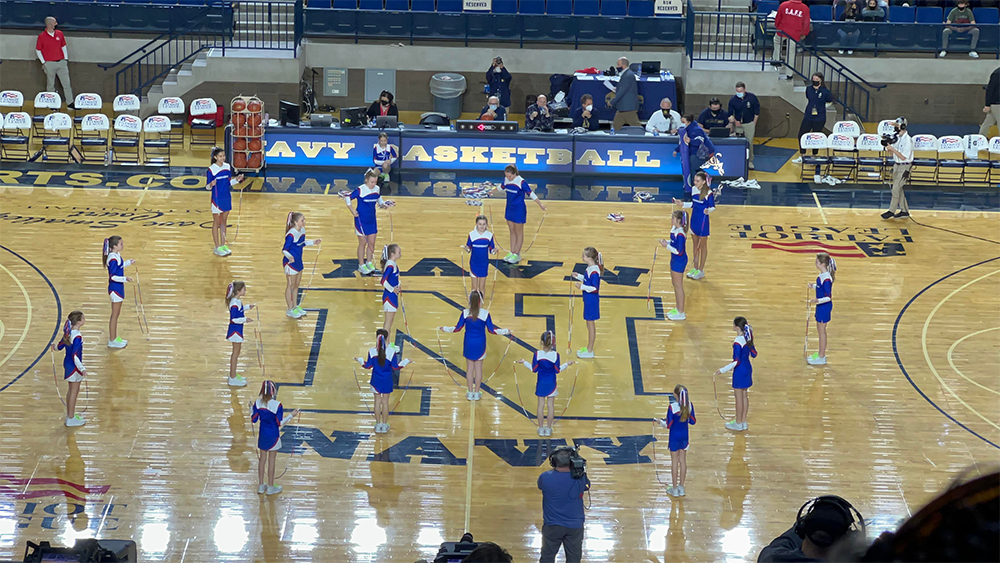 A cheerleading team in blue uniforms performs a routine at center court in a gymnasium with “NAVY” on the floor. Spectators sit in the bleachers, and some people are seated at tables on the far side of the court.