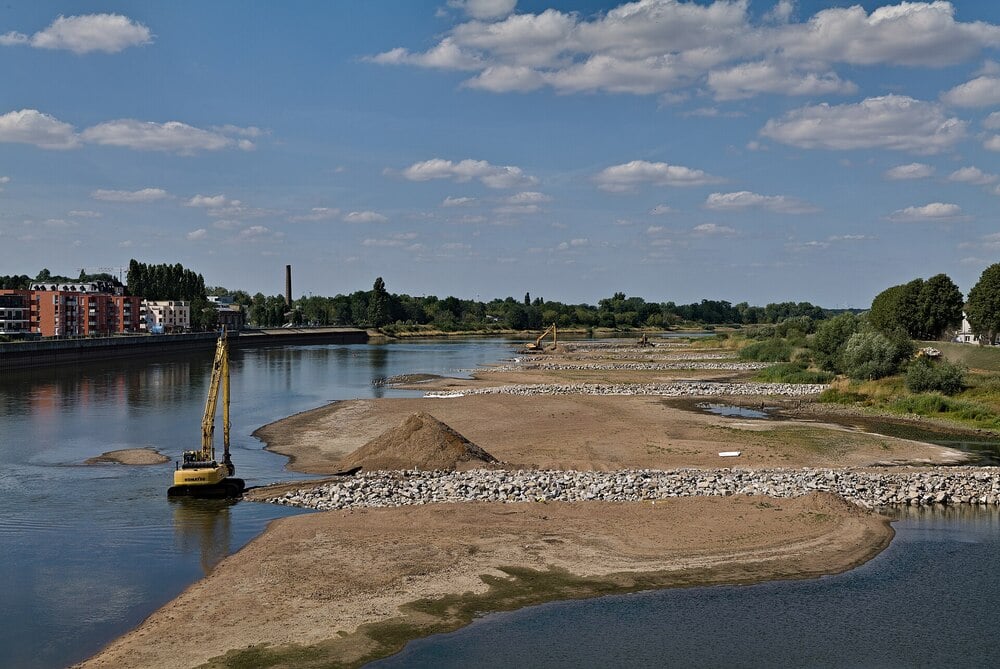 A river with exposed sandbanks, construction equipment, and rocks along the shore, surrounded by buildings, trees, and a partly cloudy blue sky.
