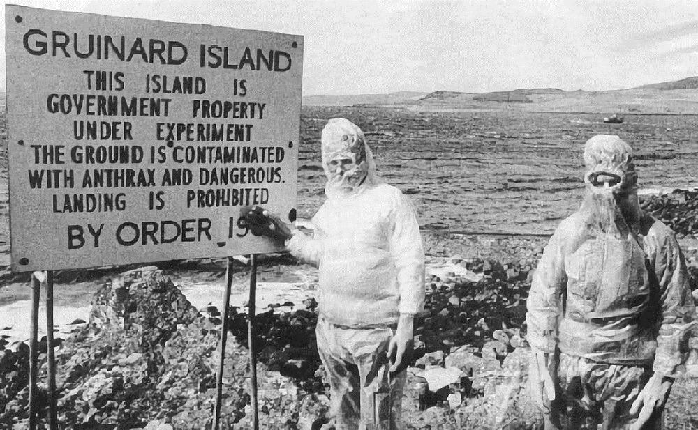 Two people in protective suits stand beside a sign warning that Gruinard Island is contaminated with anthrax and landing is prohibited. Rocky shore and water are visible in the background.