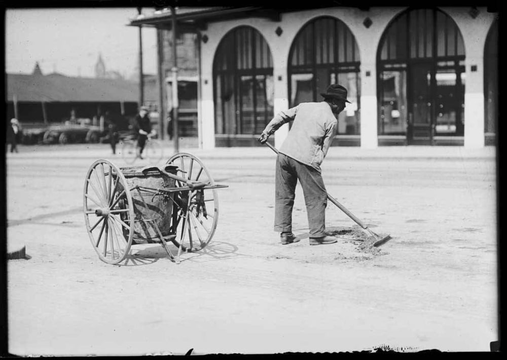 A person wearing a hat sweeps a street near a building with arched windows. A cart with large wooden wheels stands nearby, likely used for collecting debris.