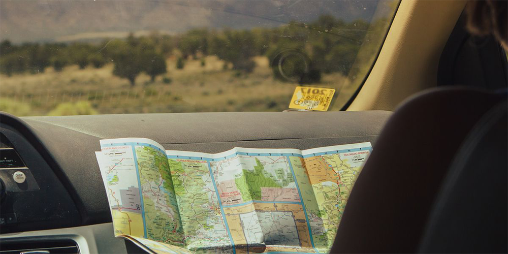 A person in a car looks at a paper map, with a landscape of trees and mountains visible through the windshield. A yellow parking permit is on the dashboard.
