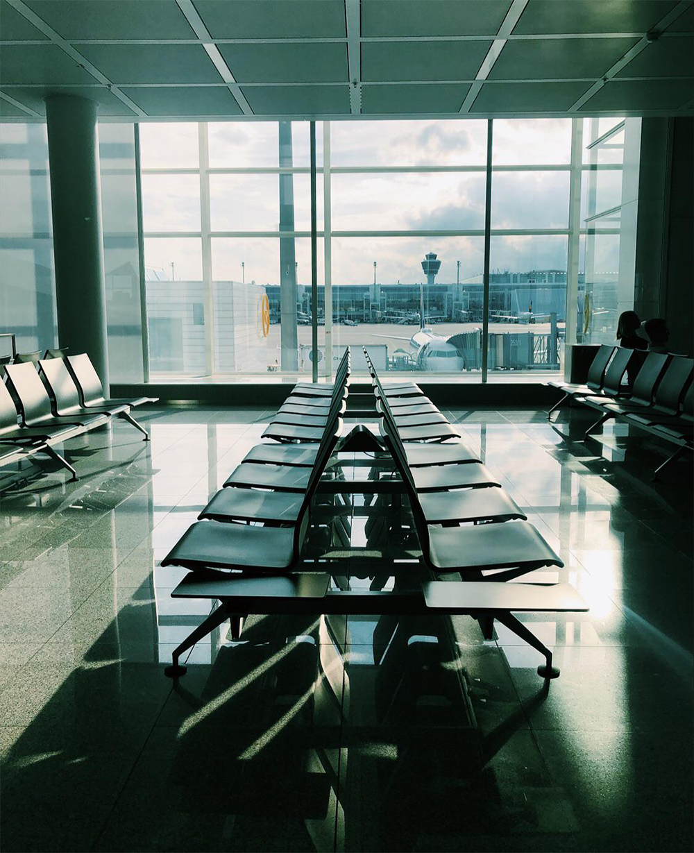 Empty airport seating area with rows of black chairs facing large windows. Outside, a plane and control tower are visible under a cloudy sky. Sunlight casts shadows on the glossy floor.