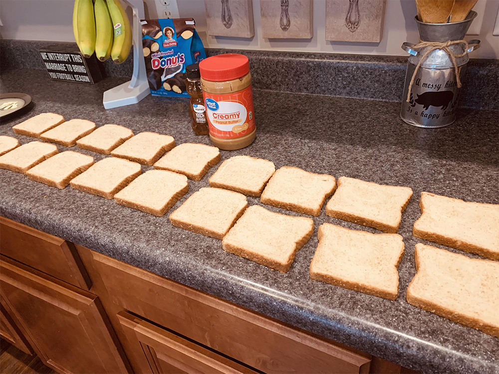 Fourteen slices of bread are lined up in pairs on a kitchen counter. Behind them are a jar of creamy peanut butter, a spice jar, Little Debbie Donut Sticks, bananas, and various kitchen utensils.