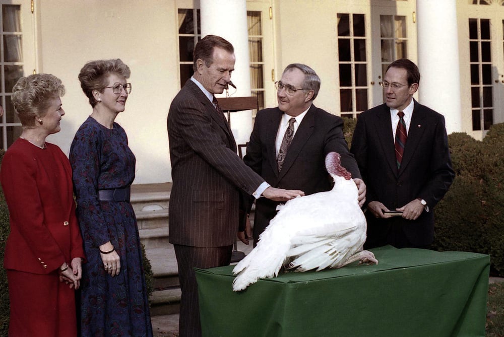 Five adults stand outside around a table with a turkey on it; one man is patting the turkey, and all are smiling. The background shows white columns and windows, suggesting a formal setting.
