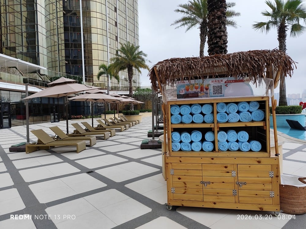 Poolside area with lounge chairs under umbrellas, a palm tree, and a wooden towel cart stocked with blue towels. A tall glass building and cloudy sky are in the background.