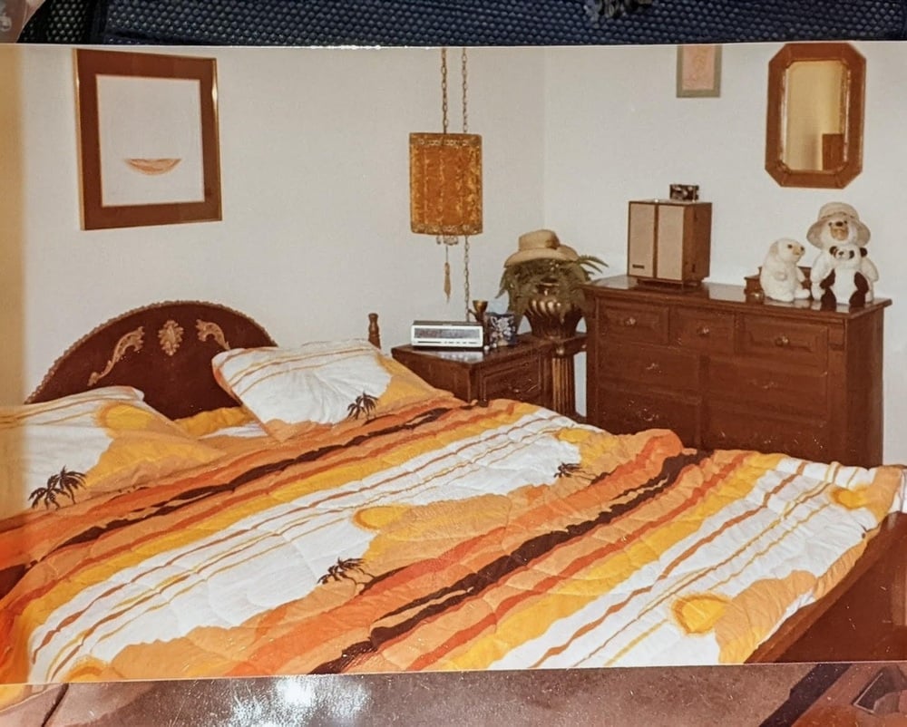 A bedroom with a yellow and orange striped bedspread, two bedside tables, a dresser with two stuffed animals, a mirror, and wall art. A hanging lamp and a potted plant are also visible.
