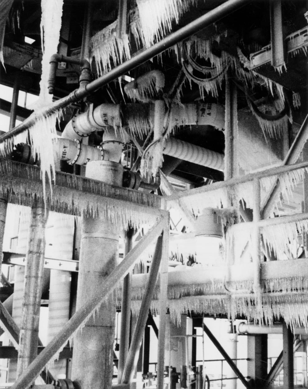 Industrial pipes and metal framework covered in long icicles and frost, indicating freezing temperatures inside a factory or processing plant.