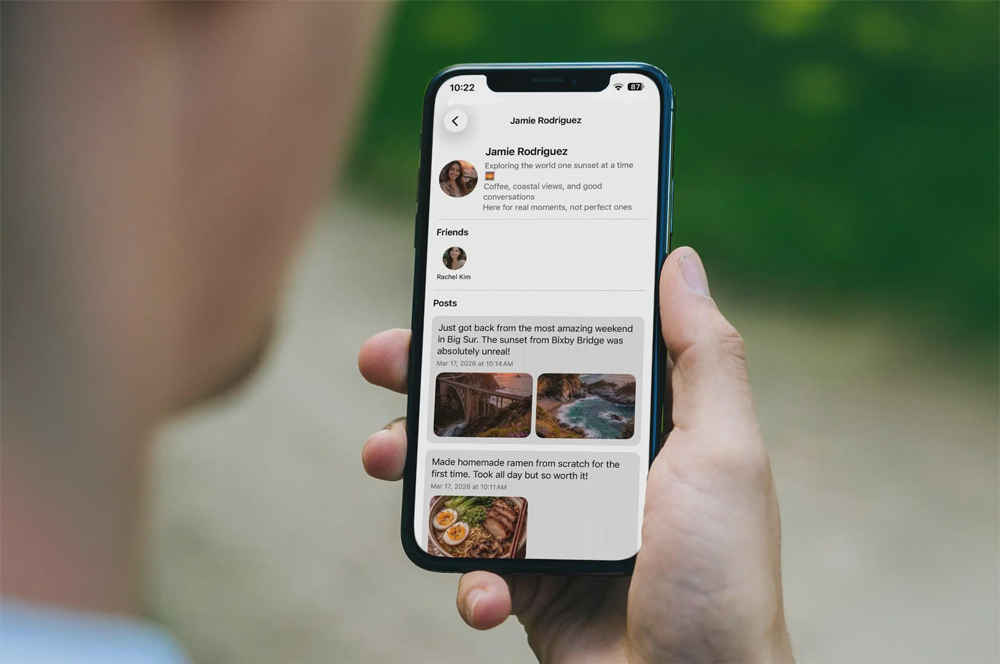A person holds a smartphone displaying a social media profile with a profile photo, bio, friends list, and posts about a weekend trip and homemade food. The phone screen is in focus, with a blurry green background.