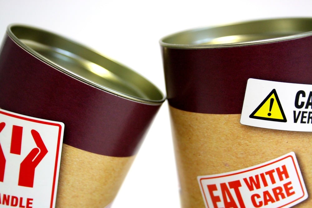 Two cylindrical cardboard containers with maroon lids. One has a “Handle with Care” sign, the other shows a warning label and reads “EAT WITH CARE.” Both are close together on a white background.