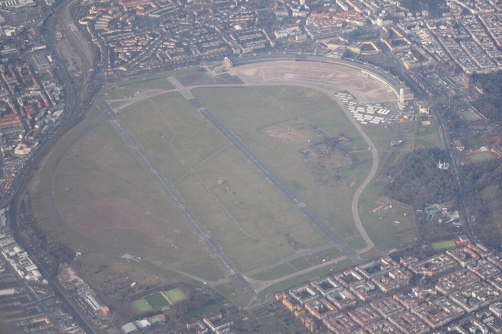 Aerial view of Tempelhofer Feld, a former airport in Berlin, with runways, grassy areas, nearby buildings, and city streets surrounding the site.