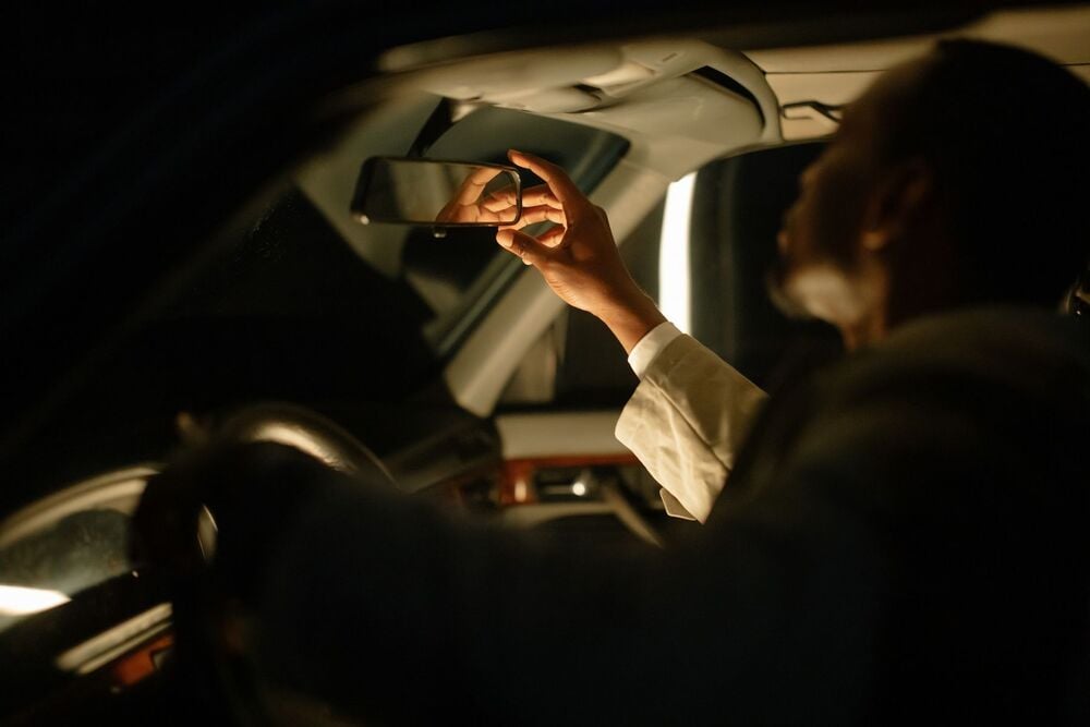 A person sitting in a car adjusts the rearview mirror at night. The interior is dimly lit, highlighting the person’s hand and face in profile.