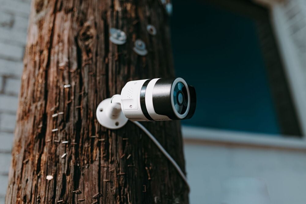 A security camera is mounted on a wooden pole covered with staples, with a blurred building in the background.