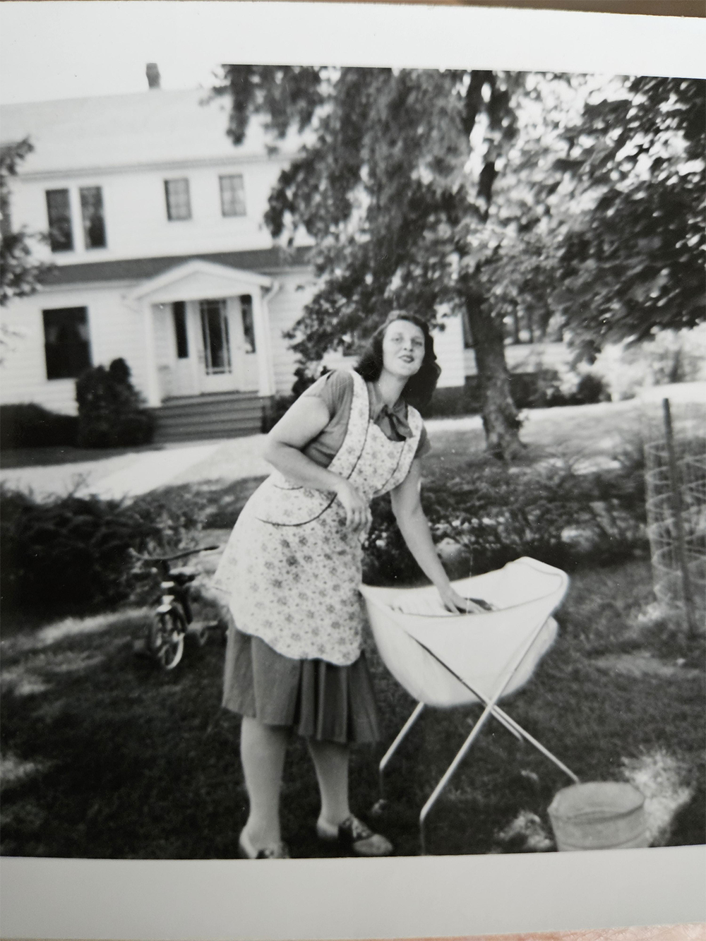 A woman in an apron stands outdoors next to a baby bassinet on a lawn, with a large house and trees in the background. The photo is black and white.