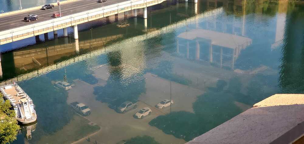 A river reflects a building and parked cars so clearly that they appear underwater, while a bridge with moving vehicles crosses above.