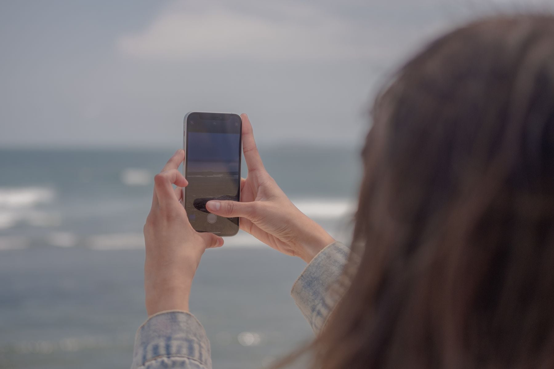 A person with long hair holds up a smartphone, taking a photo of the ocean. The sea and sky are visible in the background. The person’s face is not visible.