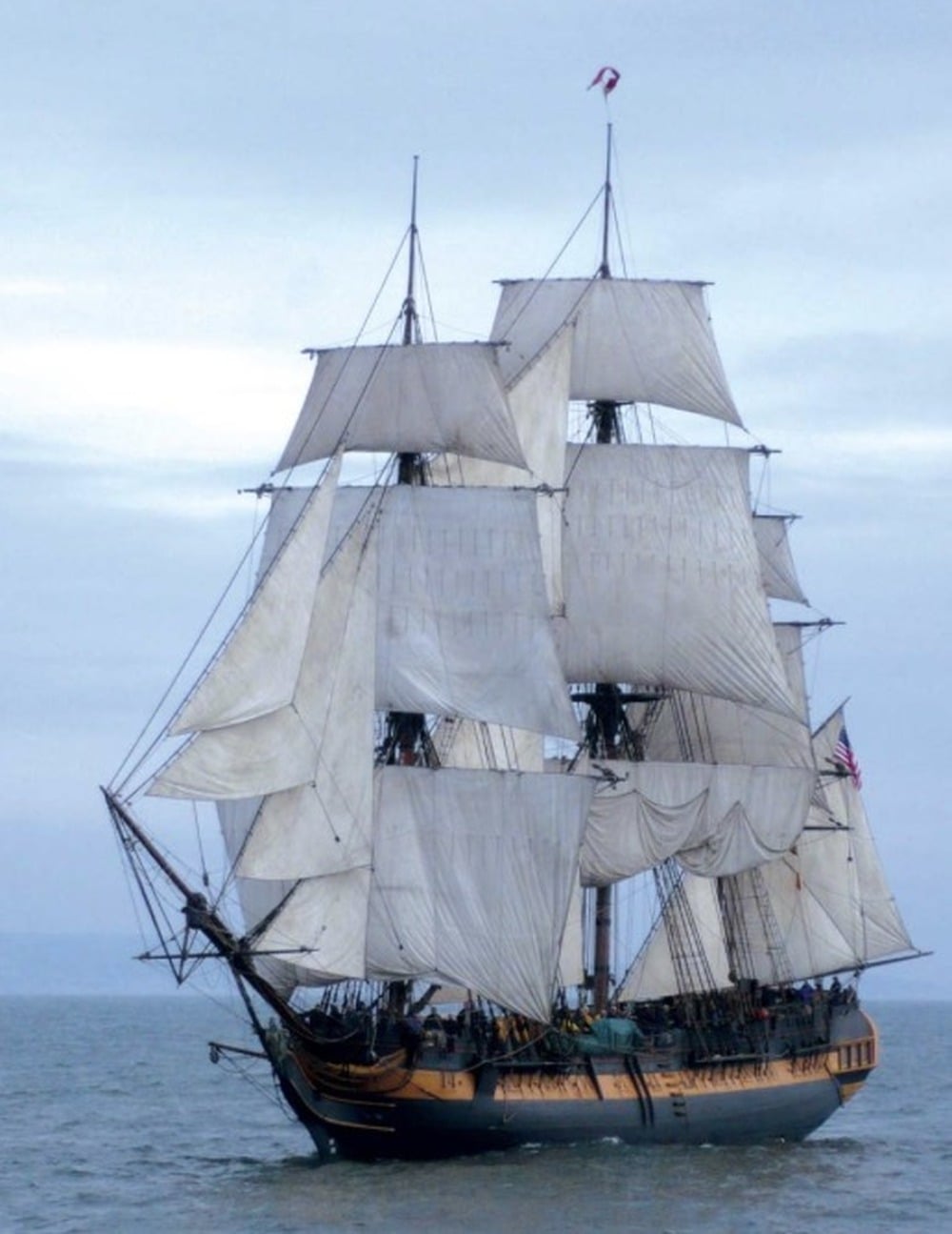 A large, historic tall ship with multiple white sails unfurled is sailing on calm waters under a cloudy sky.