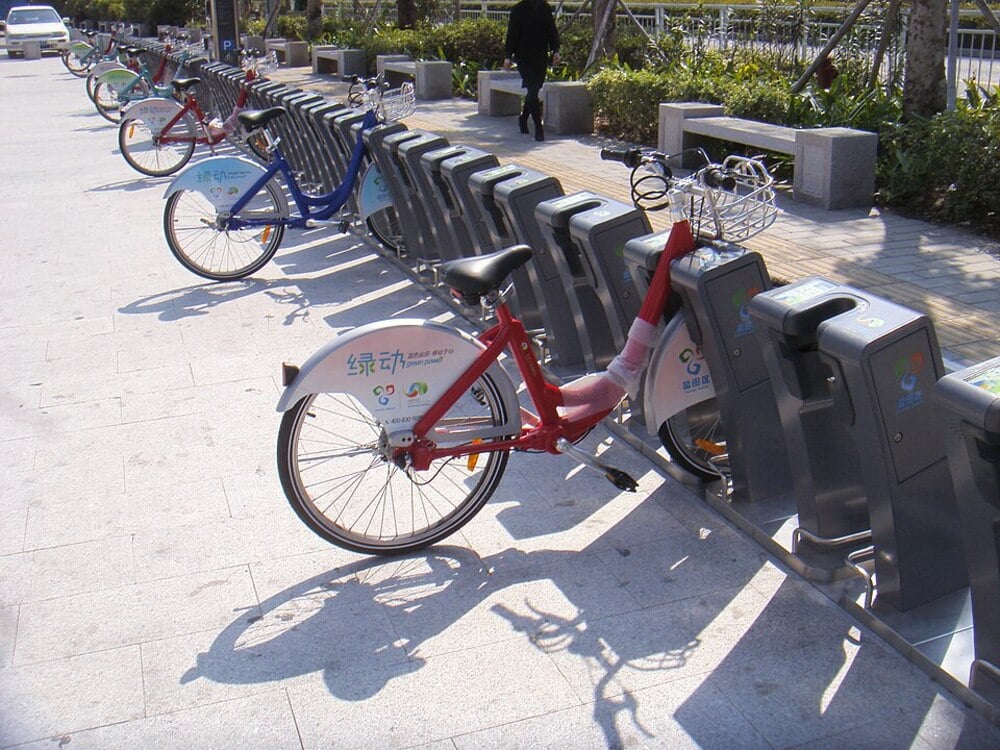 Several colorful bicycles are parked in a row at a public bike-sharing station on a sunny day, with a sidewalk, benches, greenery, and a few people in the background.