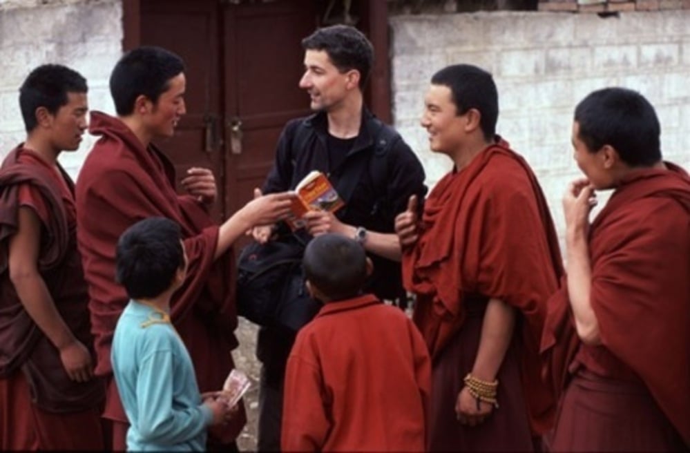 A group of monks in red robes smile and interact with a man in black clothing, who is handing them a boxed item. Two children in blue and red are also present, all appearing cheerful in an outdoor setting.