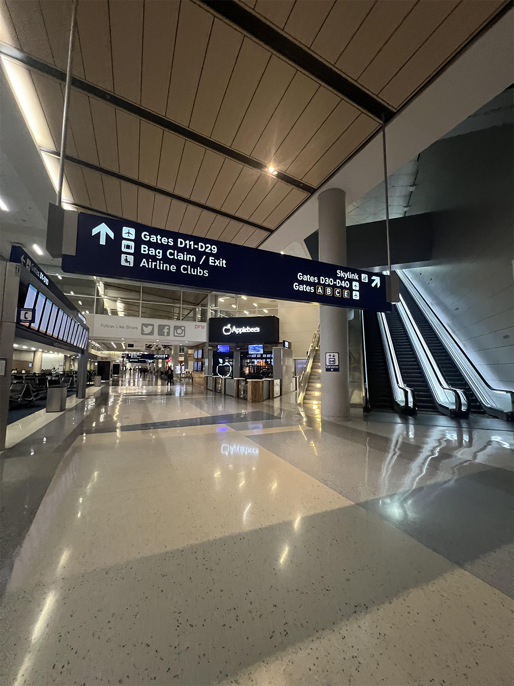 Empty airport terminal with overhead signs directing to gates, bag claim, exit, and escalators. Escalators on the right, seating areas on the left, and airport service counters in the center.