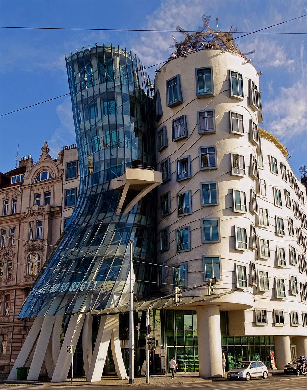 Modern building with a twisted glass tower leaning against a white, wavy structure with many windows; other traditional buildings are beside it under a blue sky.