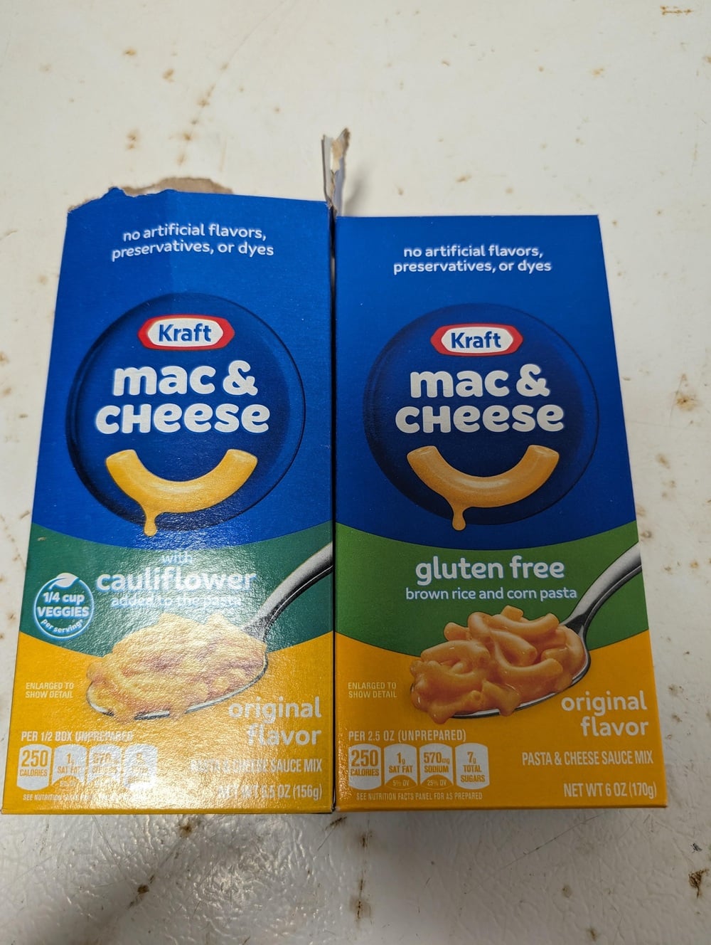 Two Kraft mac & cheese boxes on a counter: one with cauliflower, one gluten free. Both show a spoonful of pasta and have blue and yellow packaging with the Kraft logo.
