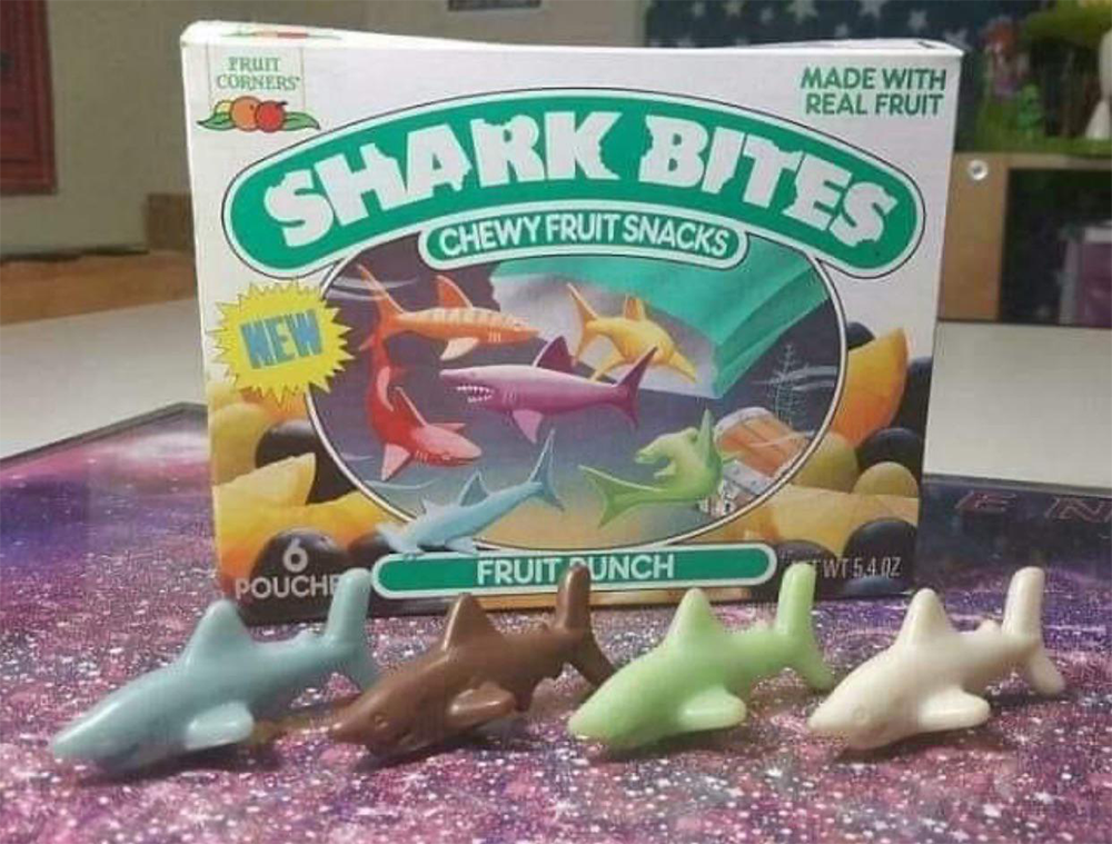 A box of Shark Bites chewy fruit snacks is shown behind four shark-shaped gummies in different colors, arranged side by side on a table.