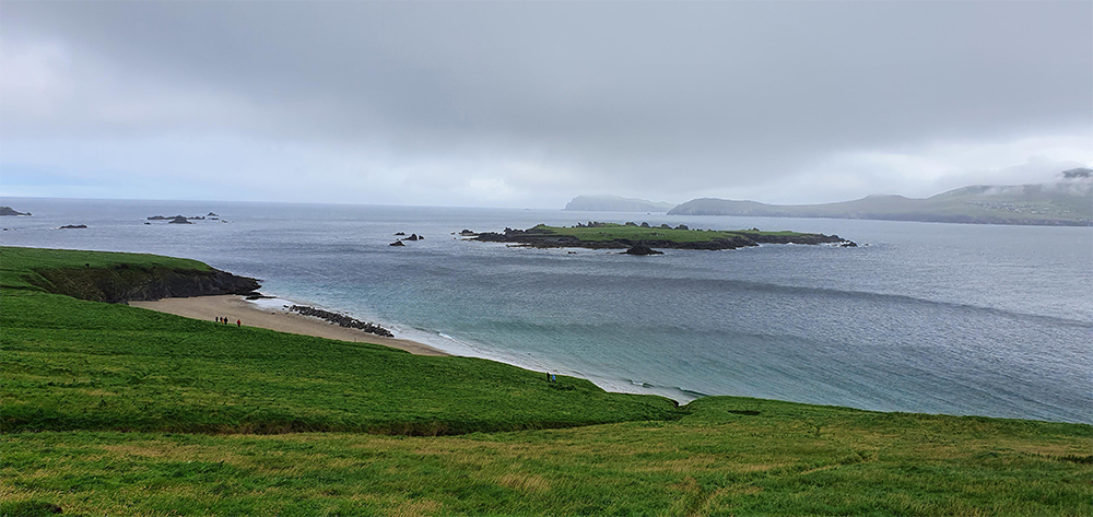 A cloudy coastal landscape with green grassy hills, a sandy beach, and rocky islands in calm blue-gray water under an overcast sky.