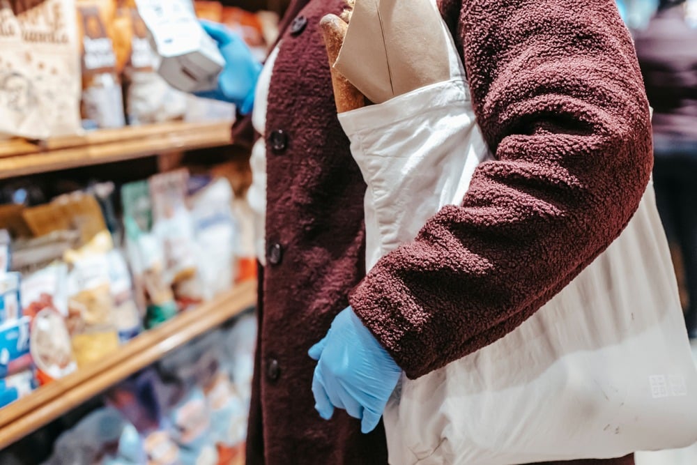 A person wearing blue gloves and a maroon coat shops in a grocery store, holding a white reusable bag with a baguette and a brown paper bag inside. Shelves with packaged goods are visible in the background.
