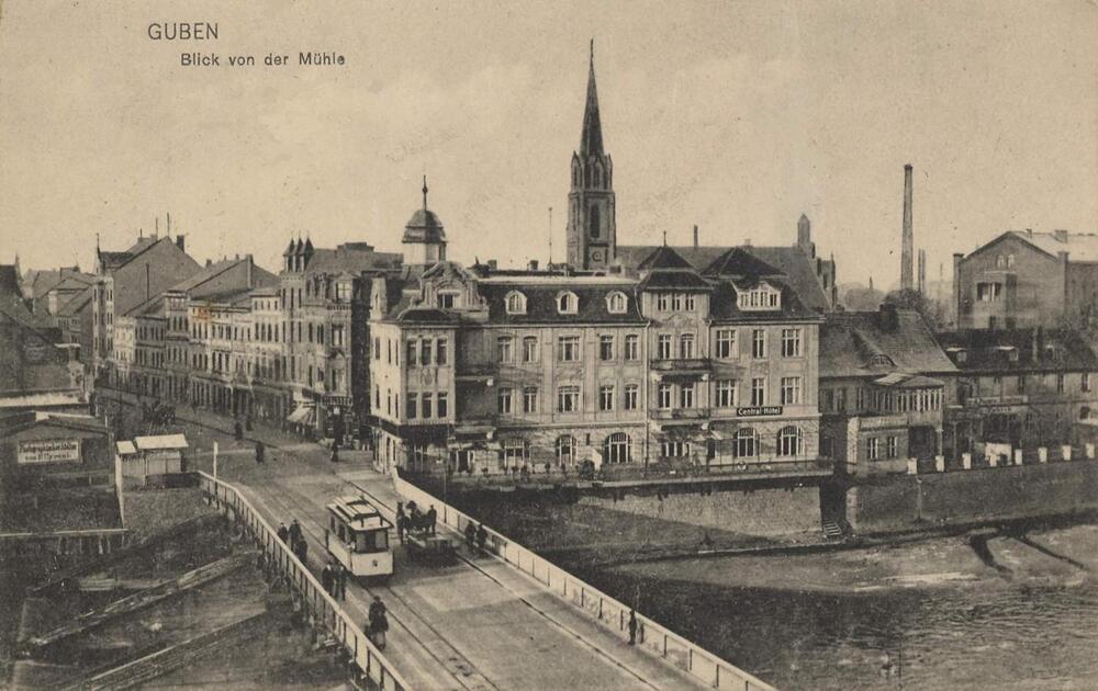 Black-and-white vintage photo of Guben, Germany, showing historic buildings, a church with a tall spire, a bridge over a river, and a tram crossing. The image is labeled “GUBEN Blick von der Mühle.”