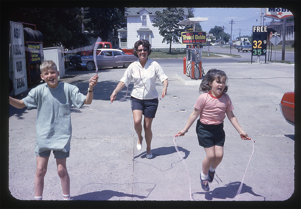 A woman and two children play jump rope outside a gas station on a sunny day. The kids are mid-jump and smiling. A gas price sign and vintage cars are visible in the background.